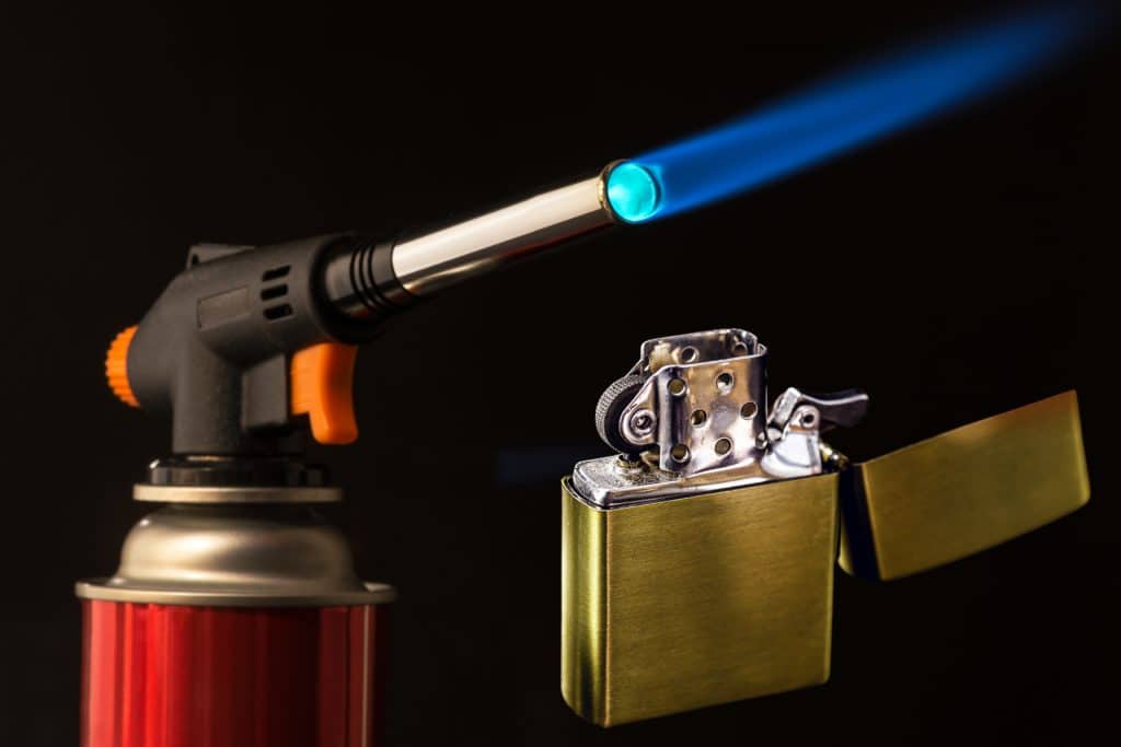 Butane torch with blue flame and red canister and gold zippo lighter in the open position 