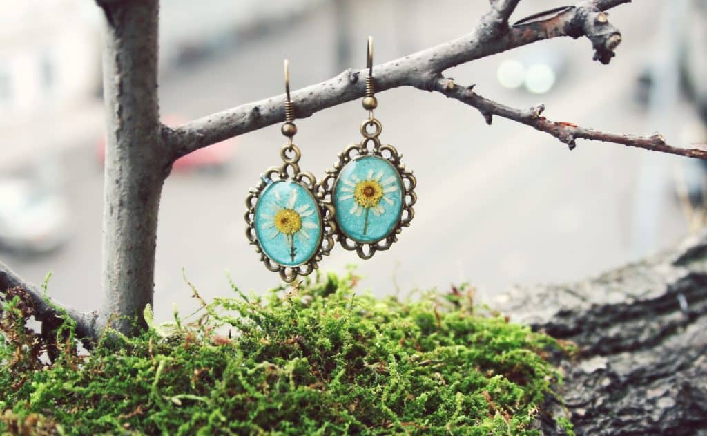 Oval shaped light blue resin earrings with an image of a sunflower inside. the earrings are hanging from a small tree branch over a small clump of moss.