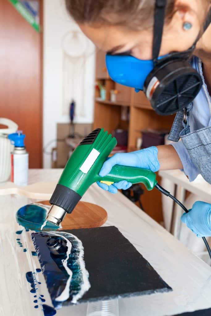 Woman with a respirator mask using a heat gun to move blue and white resin making an ocean wave effect 