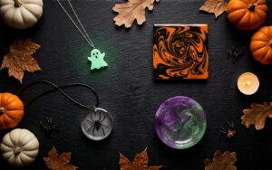 Finished Halloween resin crafts styled together on black slate — a glow-in-the-dark ghost pendant, black and orange marbled coaster, spider embed pendant and purple-green witch's-brew trinket dish