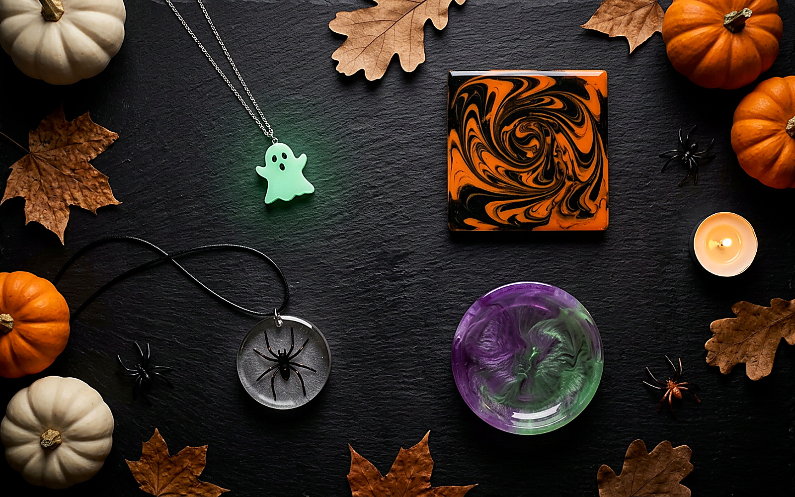 Finished Halloween resin crafts styled together on black slate — a glow-in-the-dark ghost pendant, black and orange marbled coaster, spider embed pendant and purple-green witch's-brew trinket dish