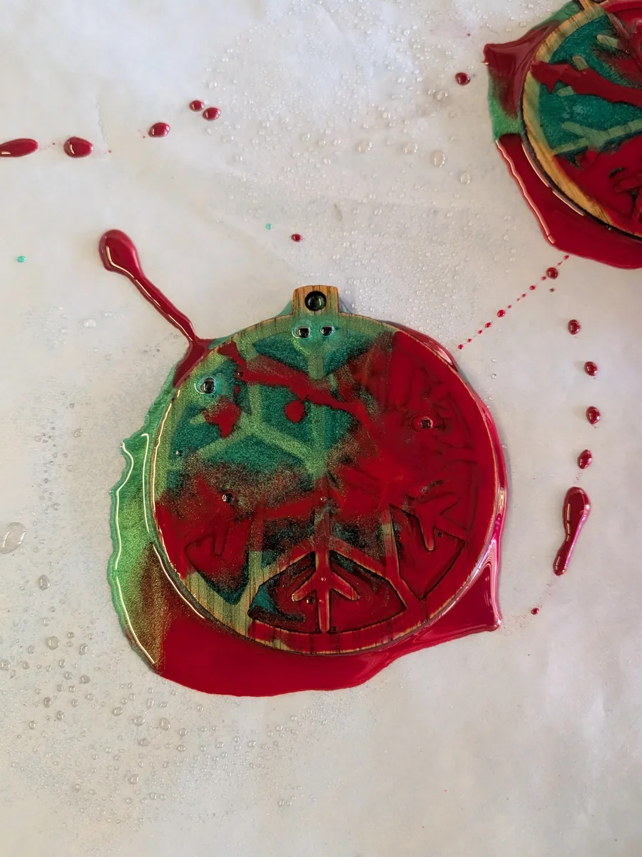 Red and green epoxy ornament with resin overflowing its edges and pooling on the paper underneath