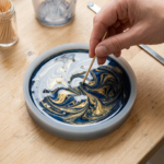 A hand swirling a toothpick through wet navy blue and gold pigmented epoxy resin inside a round silicone trinket dish mold, creating a marbled swirl pattern.
