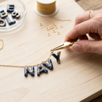 Navy-and-gold resin letters being linked with jump rings and pliers onto a gold chain to form a name necklace, on a wooden workbench.