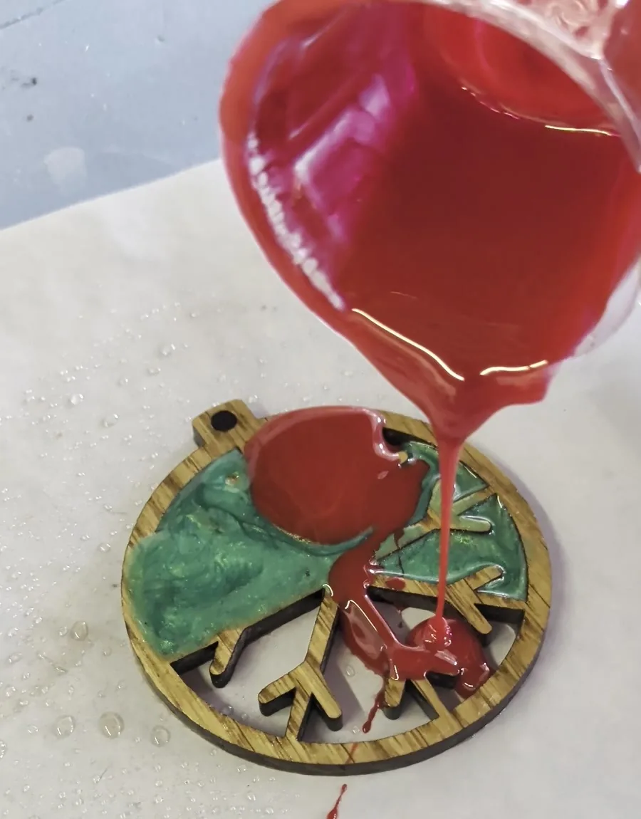Red-tinted epoxy pouring from a small cup in a steady stream onto a laser-cut wooden snowflake ornament