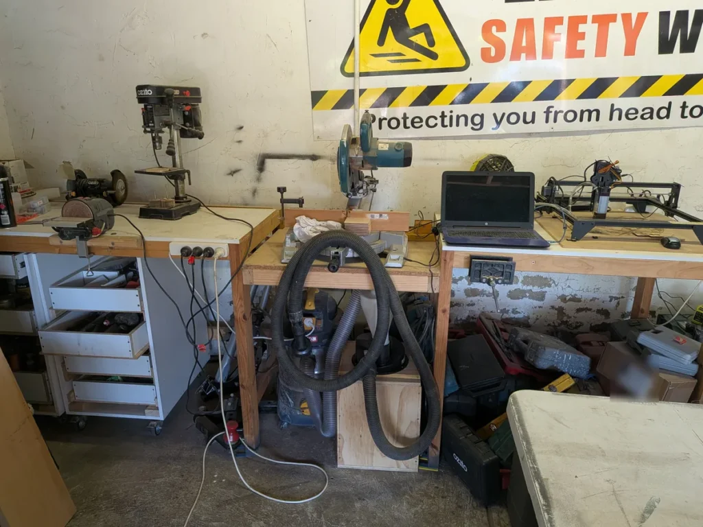 A double-garage workshop with wooden benches holding a drill press, bench grinder, miter saw and a small laser engraver, with a shop vac hose and dust separator under the bench