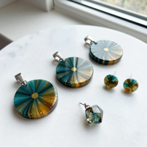 A flat lay of resin jewelry for beginners — three round pendants and a pair of stud earrings in swirled teal and amber-gold with gold mica flecks, plus a faceted geodesic charm, on white marble near a window