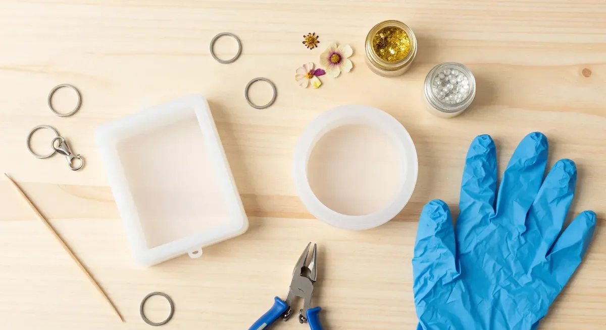 Resin keychain supplies laid out on a wooden table: silicone keychain molds, split key rings, a lobster clasp, dried flowers, jars of gold glitter and pearls, pliers, a toothpick and a nitrile glove