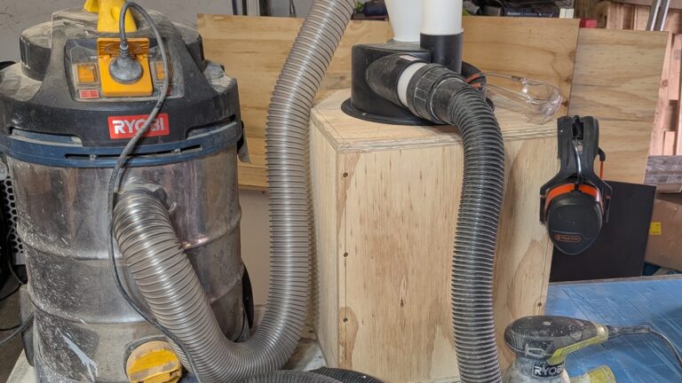 Old Ryobi shop vac connected by hose to a cyclone dust separator on a plywood bin, with ear muffs and a random orbital sander