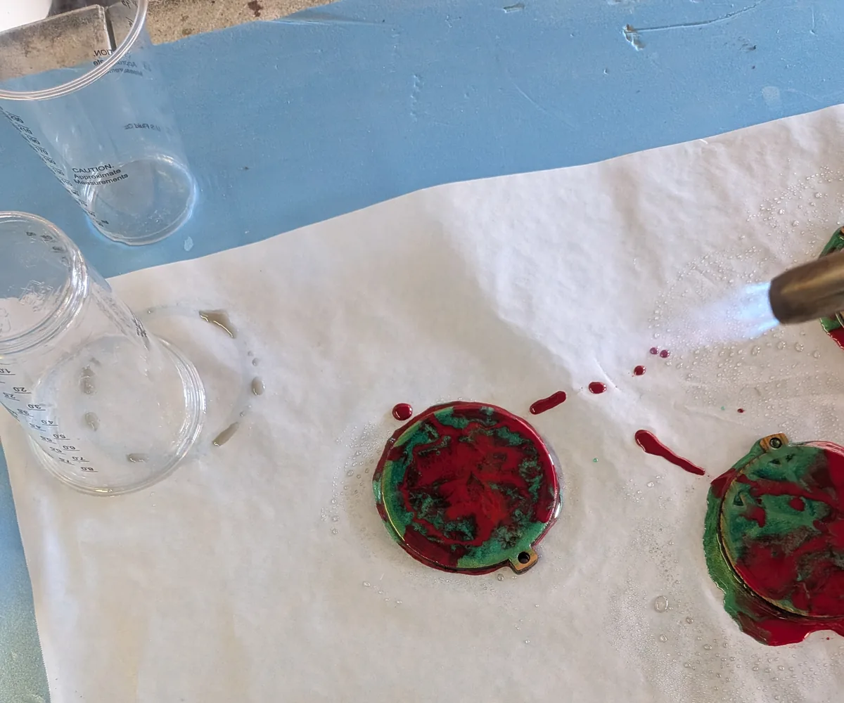 Butane torch passing over freshly poured red and green epoxy ornaments on paper