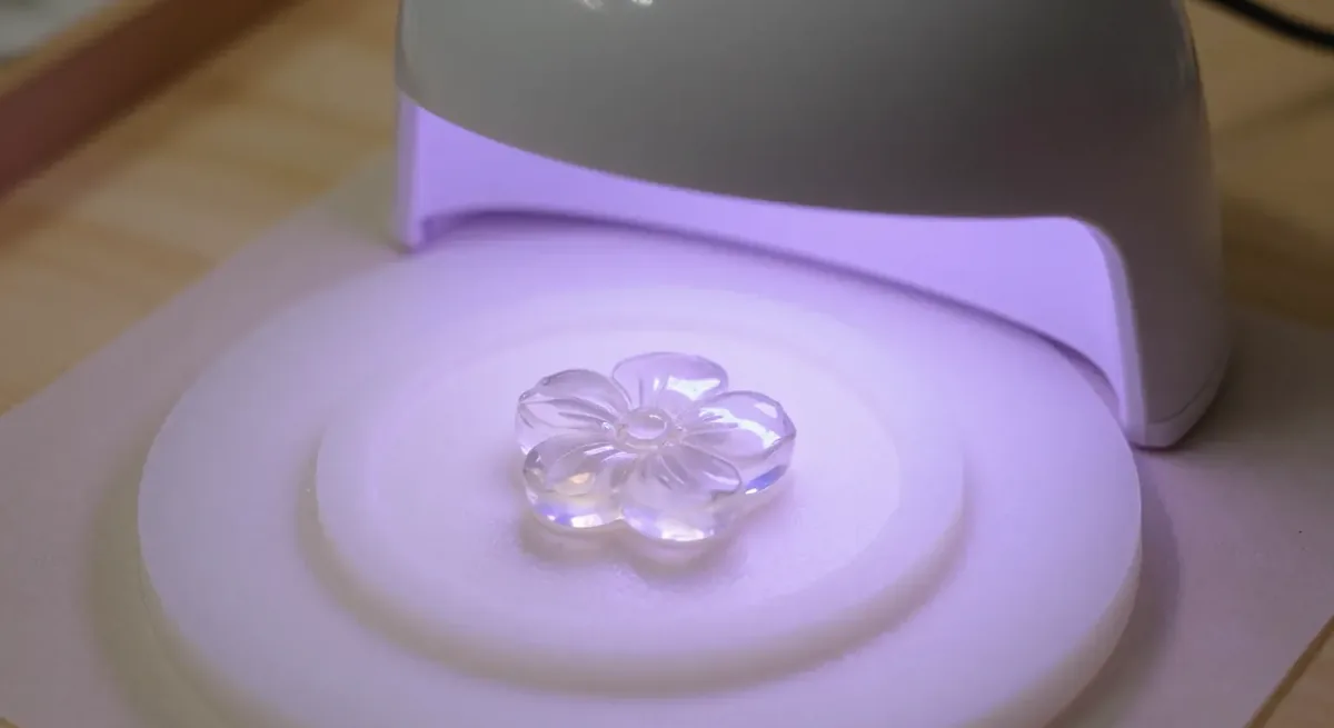 A clear resin flower pendant curing under a small dome-shaped UV lamp with a soft violet glow