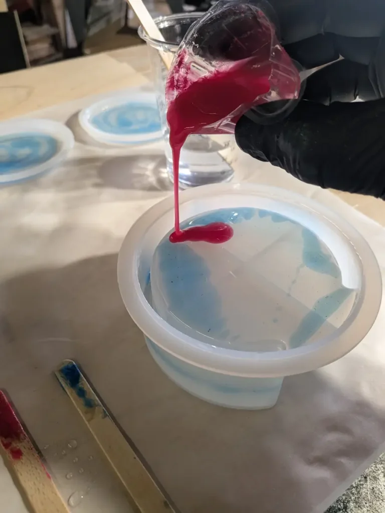 A gloved hand pouring red-tinted epoxy resin into a deep round silicone mold that already holds blue resin