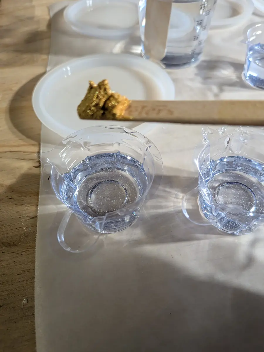 Gold mica paste on a wooden stir stick held over small cups of clear mixed epoxy resin