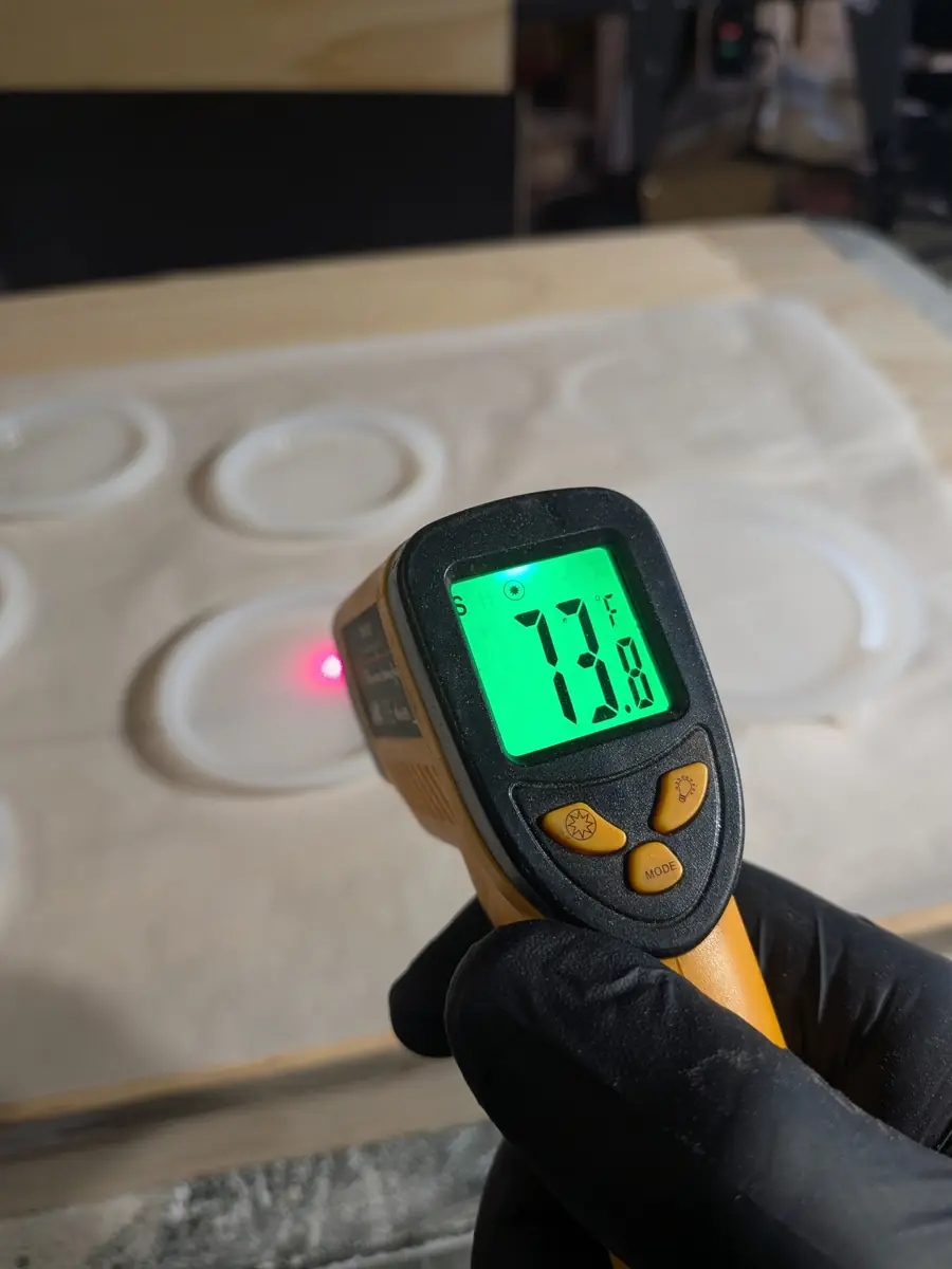 A gloved hand holding an infrared thermometer reading 73.8°F at the coaster molds before the pour