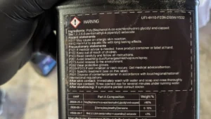Gloved hand holding a Let's Resin epoxy Part A bottle, its back label showing a WARNING signal word and hazard statements and no food-contact claim