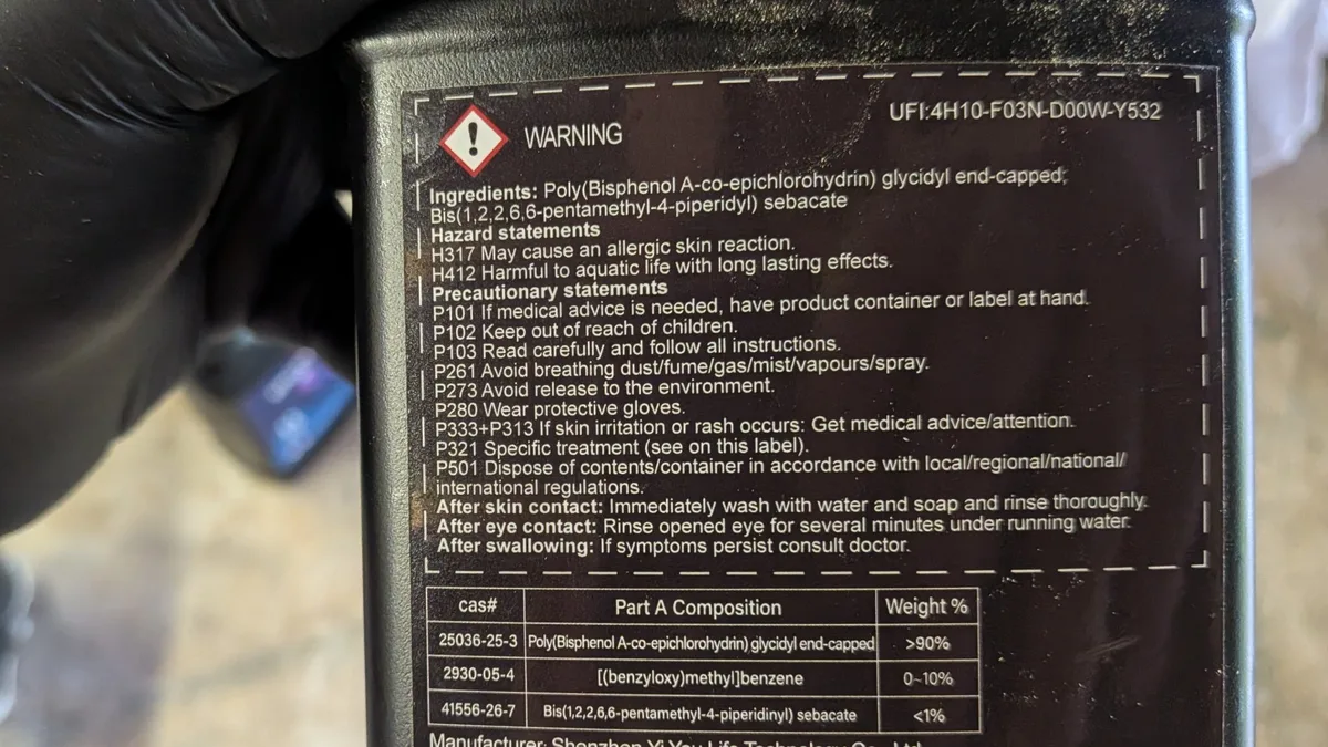 Gloved hand holding a Let's Resin epoxy Part A bottle, its back label showing a WARNING signal word and hazard statements and no food-contact claim