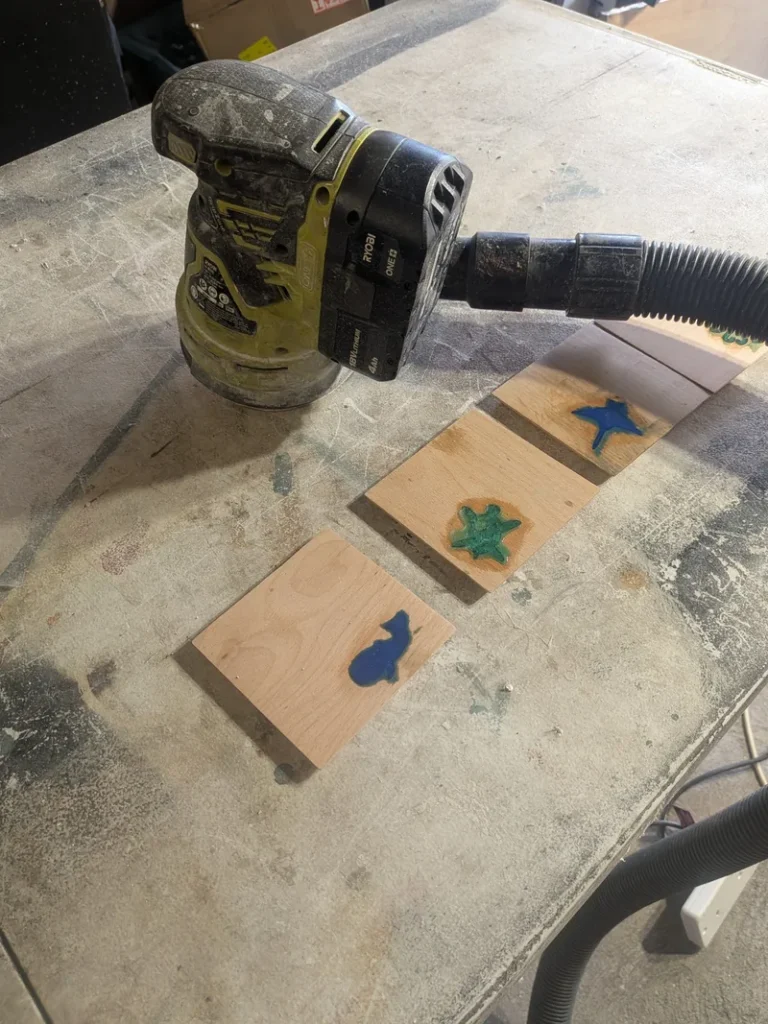 Random orbit sander with a vacuum hose fitted, next to four resin-filled beech coasters before sanding