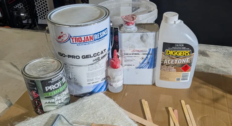 Polyester fiberglass resin, white gelcoat (also a polyester product), wax-and-styrene additive, MEKP catalyst and acetone on a workbench beside chopped strand mat