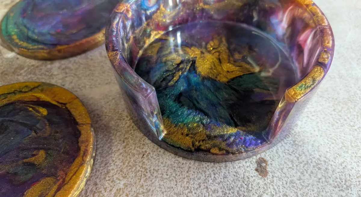 Close-up of a cured resin coaster holder with gold, teal and purple swirls, beside two finished coasters