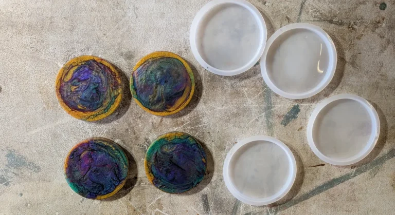 Four purple, gold and teal epoxy resin coasters laid out on a workbench beside four empty round silicone coaster molds