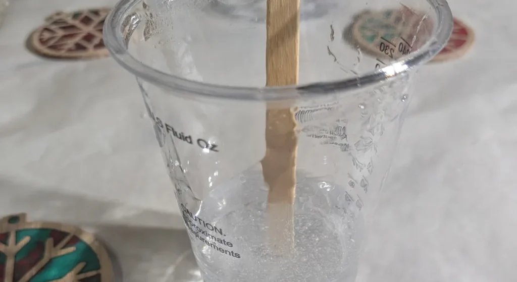 A clear mixing cup of freshly stirred epoxy resin, cloudy with fine bubbles, with a wooden stir stick standing in it