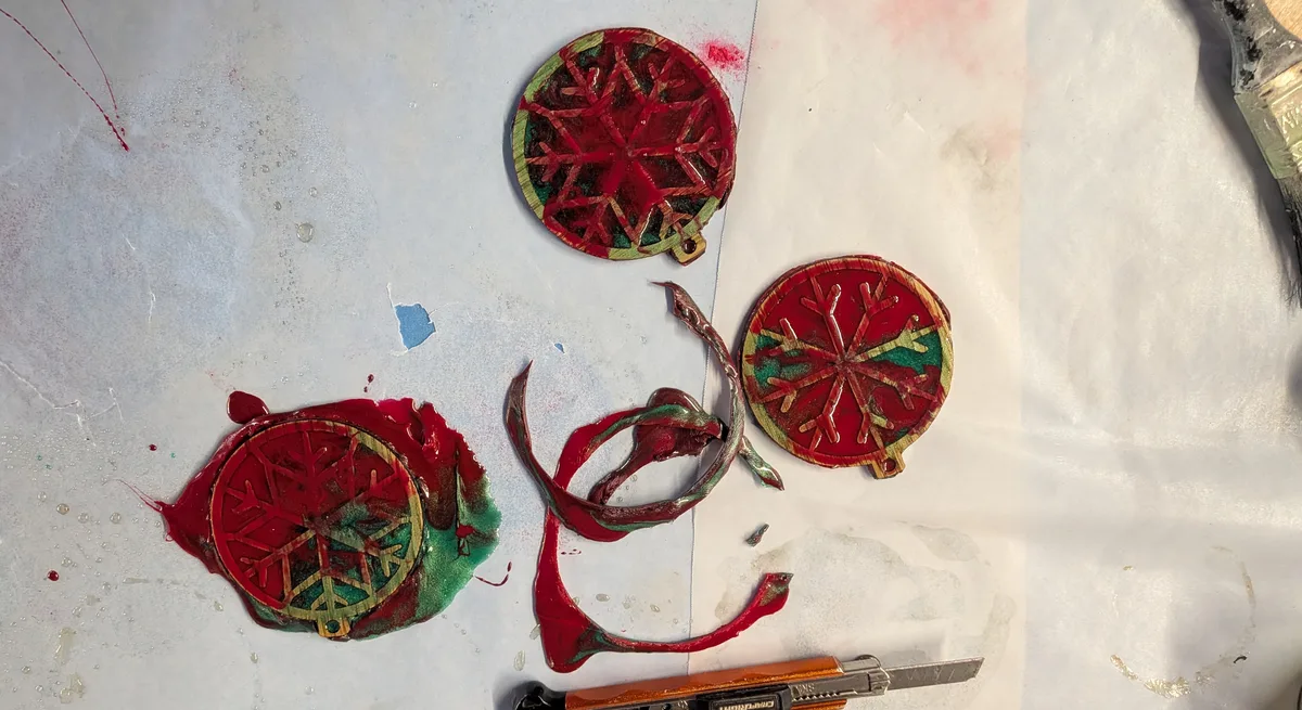 Three wood and resin ornaments on a bench, two with the rubbery red runoff cut away in strips, a craft knife beside them