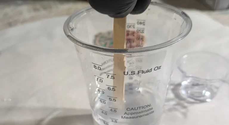 A hand in a black nitrile glove stirring clear epoxy resin in a graduated mixing cup with a wooden stick